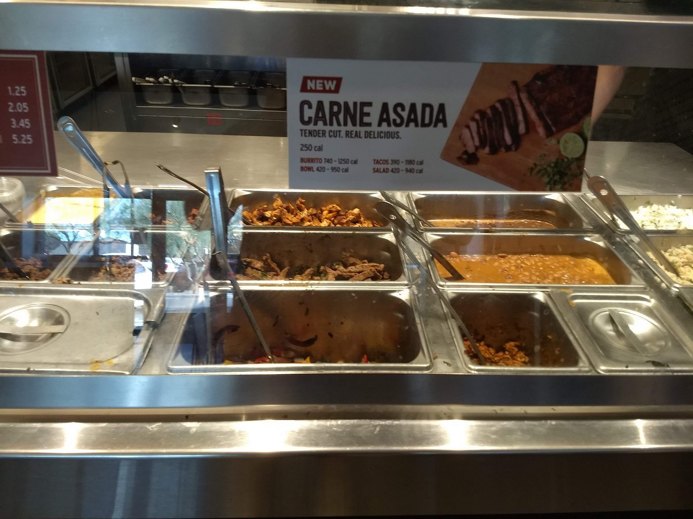 A metal Chipotle food counter displays various hot foods in trays, including meats, beans, and rice. Above, a sign advertises "Carne Asada: Tender Cut. Real Delicious." with pricing and a photo of sliced steak.