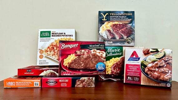 A variety of frozen meatloaf meal boxes are displayed on a wooden surface, including brands like Healthy Choice, Banquet, Marie Callender’s, Lean Cuisine, and Atkins, all with images of meatloaf and sides on the packaging.