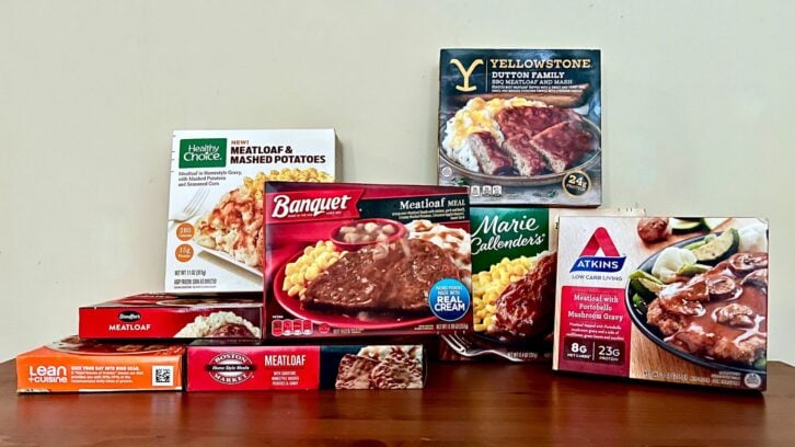 A variety of frozen meatloaf meal boxes are displayed on a wooden surface, including brands like Healthy Choice, Banquet, Marie Callender’s, Lean Cuisine, and Atkins, all with images of meatloaf and sides on the packaging.