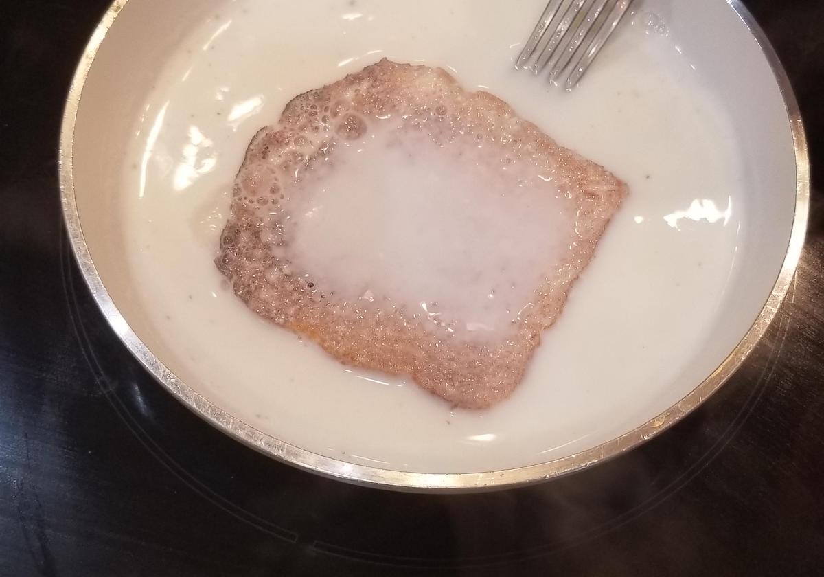 A slice of bread is soaking in a bowl of milk, with a fork resting at the edge of the bowl. Bubbles are visible on the surface of the milk around the bread.