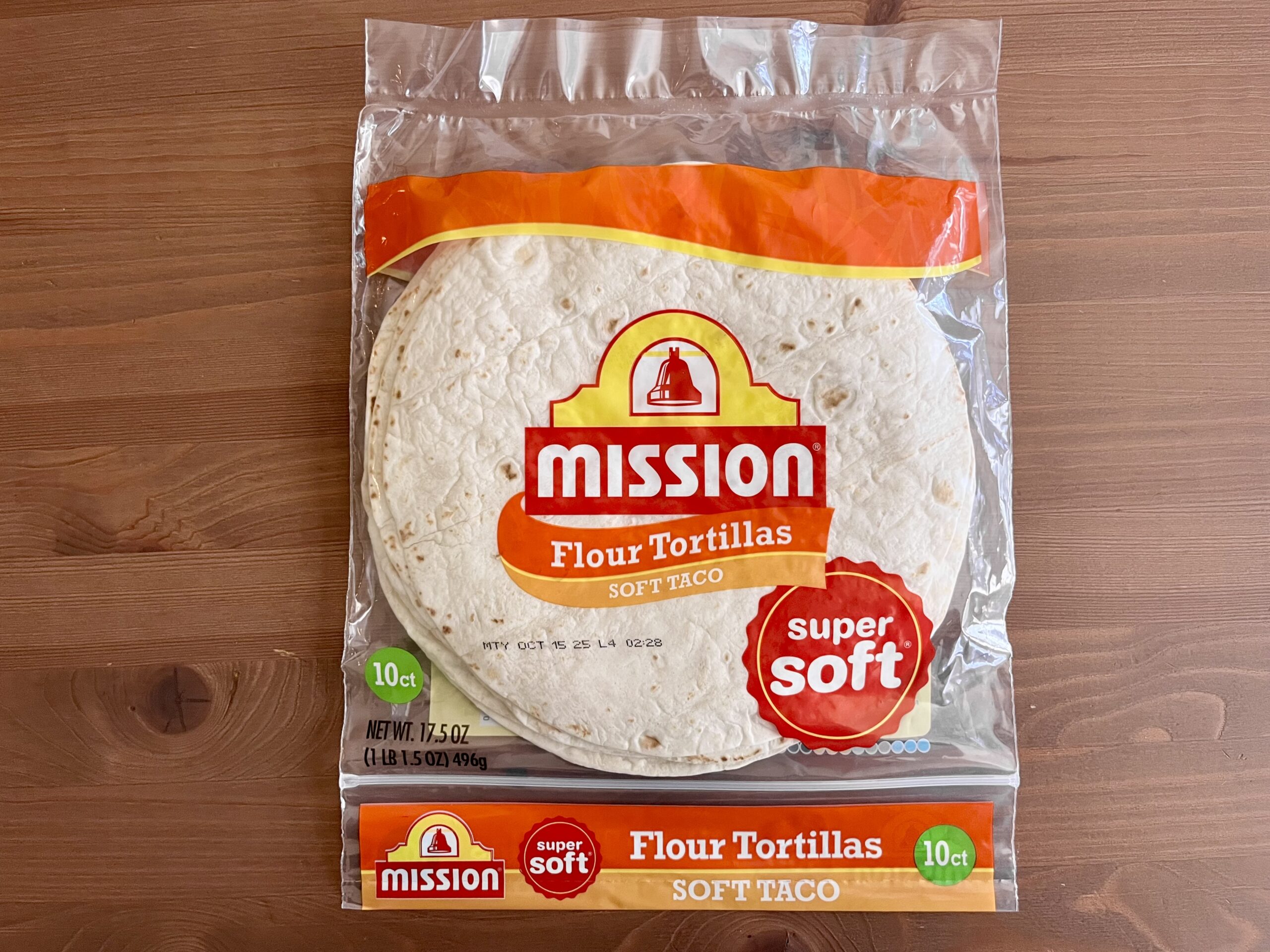 A package of Mission brand super soft flour tortillas for soft tacos sits on a wooden surface. The clear bag shows the tortillas inside and displays bright orange and yellow branding. The package contains 10 tortillas.