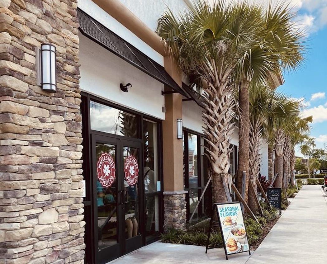 The image shows the exterior of a restaurant with stone accents, glass doors featuring a red logo, palm trees along the sidewalk, and a sign advertising seasonal flavors near the entrance. The sky is blue with some clouds.