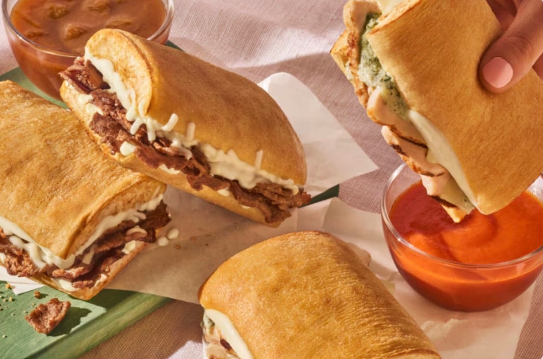 Four toasted sandwiches with melted cheese and meat fillings are arranged on a table, new fast-food style, with two bowls of dipping sauces nearby. A hand is holding one sandwich, dipping it into a bowl of red sauce.