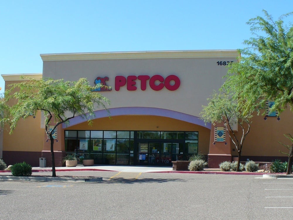 A Petco store with large red letters above the entrance, surrounded by trees and a mostly empty parking lot on a clear, sunny day.