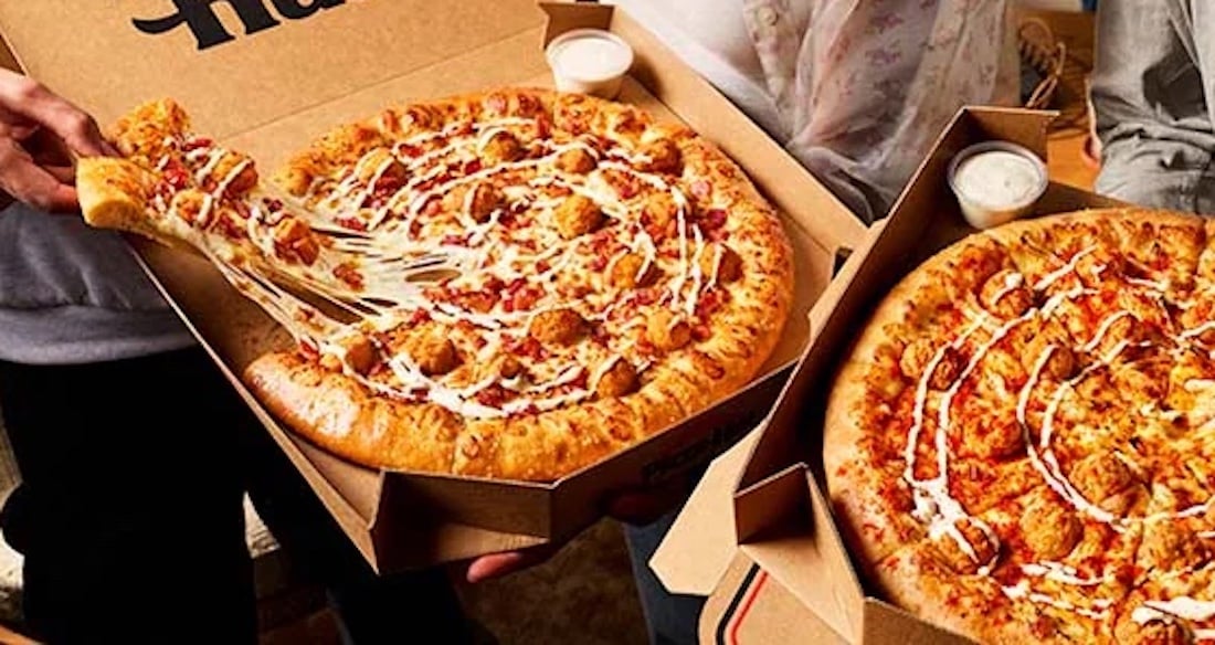 Two people holding open pizza boxes from a new fast-food spot. Each pizza has thick crust, melted cheese, chicken pieces, and a white sauce drizzle. One person pulls a cheesy slice while cups of dipping sauce sit in each box.