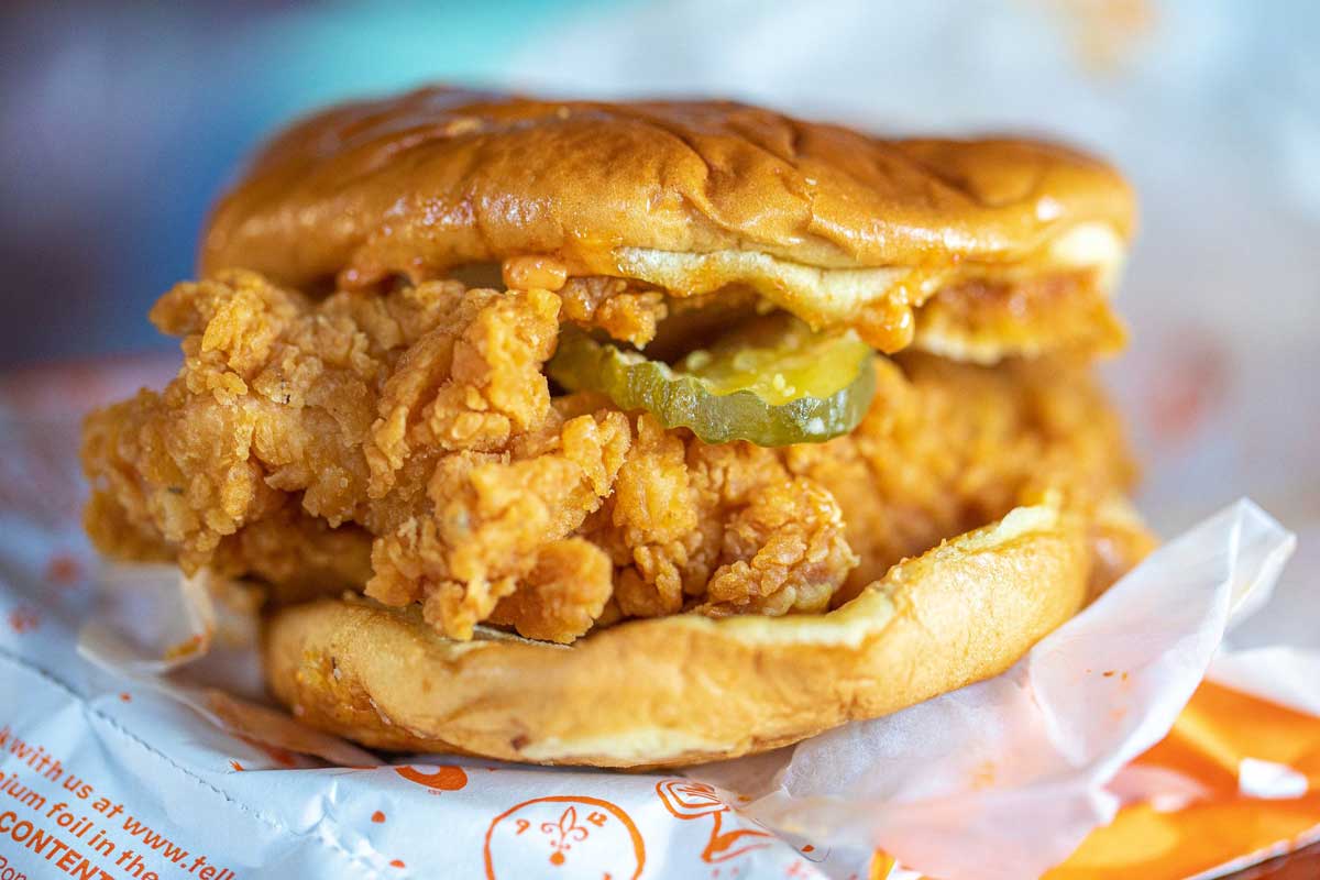 A close-up of a fried chicken sandwich with pickles, served on a soft, toasted bun—reminiscent of classic fast-food breakfast sandwiches—wrapped partly in white paper with orange print.