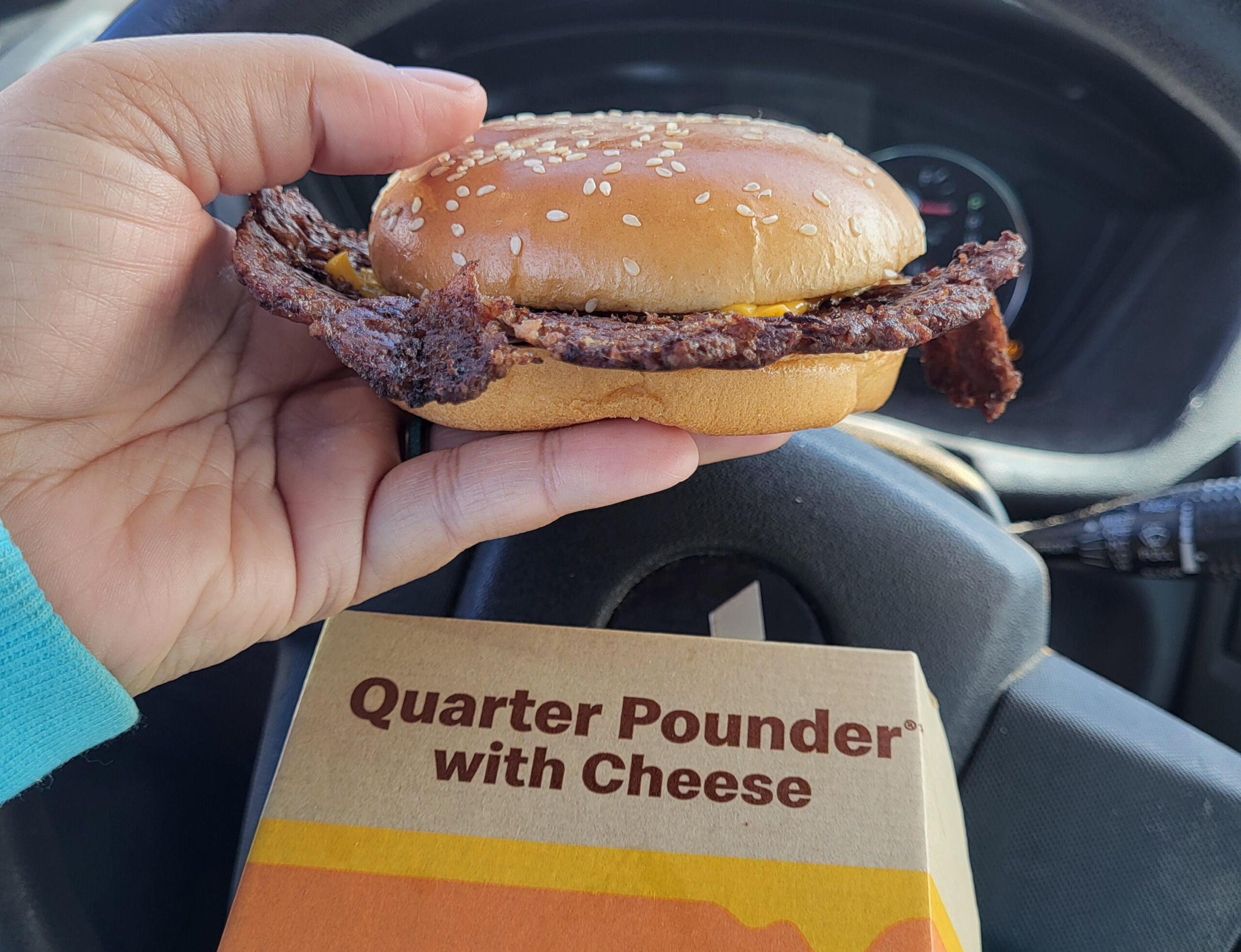 A hand is holding a Quarter Pounder with Cheese burger, showing overcooked beef edges sticking out from the bun. The steering wheel and dashboard are visible in the background, with the burger box below.