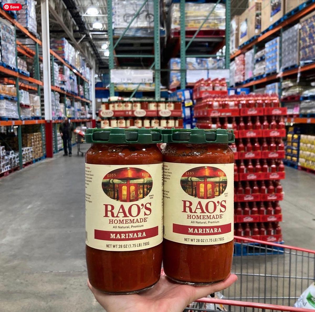 A hand holds two large jars of Rao’s Homemade Marinara sauce—one of the Costco products loved by shoppers—in a warehouse store aisle, with shelves stocked with various goods in the background.
