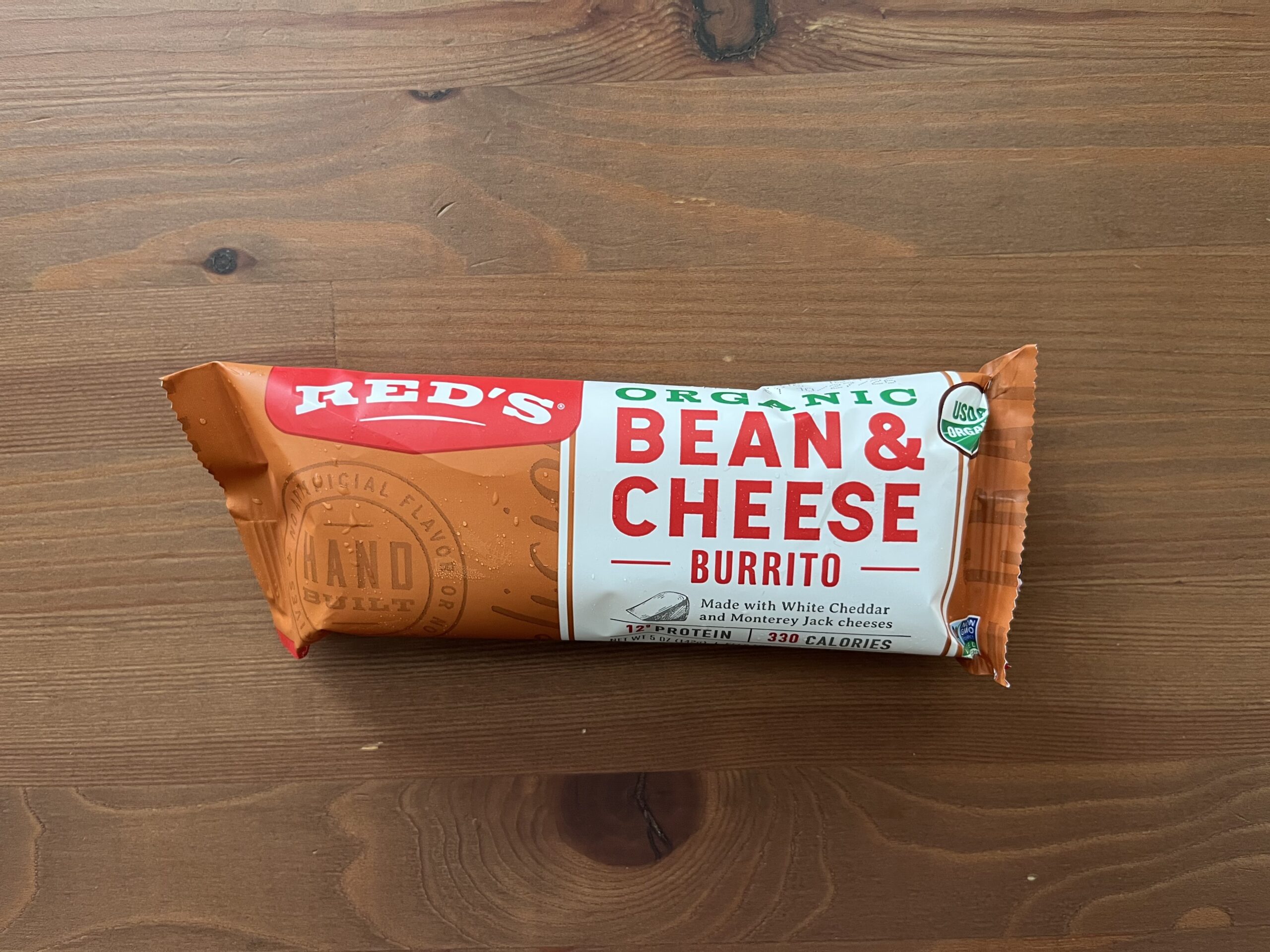 A packaged Red’s Organic Bean & Cheese Burrito sits on a wooden surface. The wrapper is orange and red, showing that it contains white cheddar and Monterey Jack cheeses, with 11g protein and 330 calories.