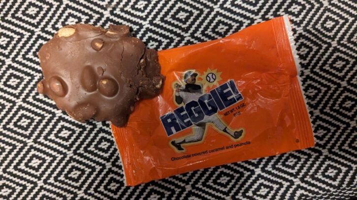 A chocolate-covered caramel and peanut candy sits next to its orange wrapper labeled "REGGIE!" with an illustration of a baseball player, on a black and white geometric patterned surface.