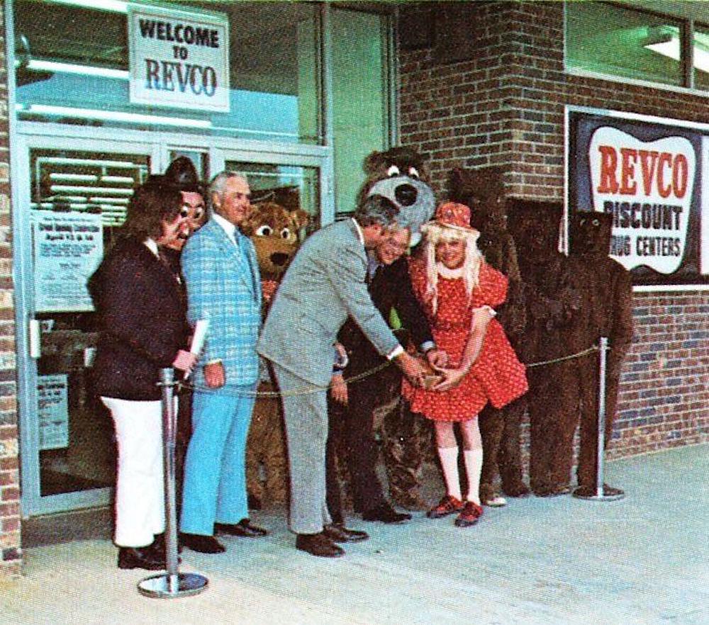 A group of people, some in animal costumes and one dressed as a doll, stand outside a Revco Discount Drug Center for a ribbon-cutting ceremony under a "Welcome to Revco" sign.