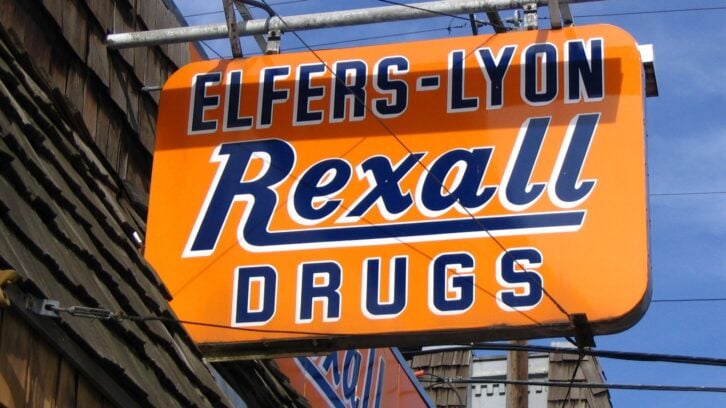 An orange and blue sign reads "ELFERS-LYON Rexall DRUGS" against a partly cloudy sky, mounted above a shingled building.