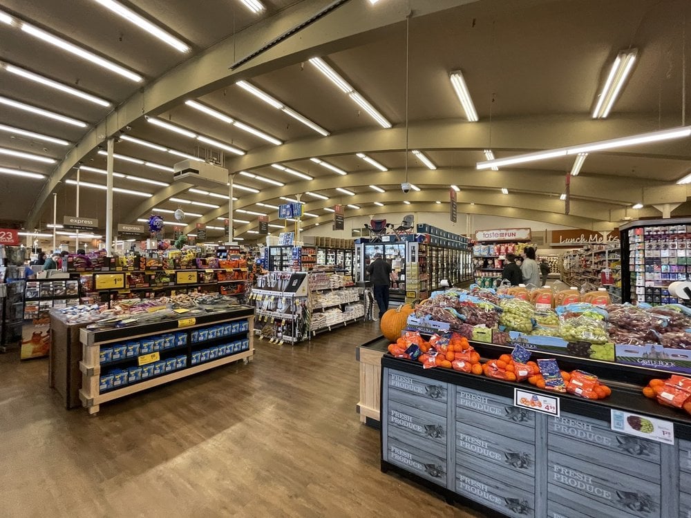 Wide-angle view of a grocery store interior with fresh produce displays in the foreground, shelves stocked with various products, and shoppers browsing the aisles under bright, curved ceiling lights.