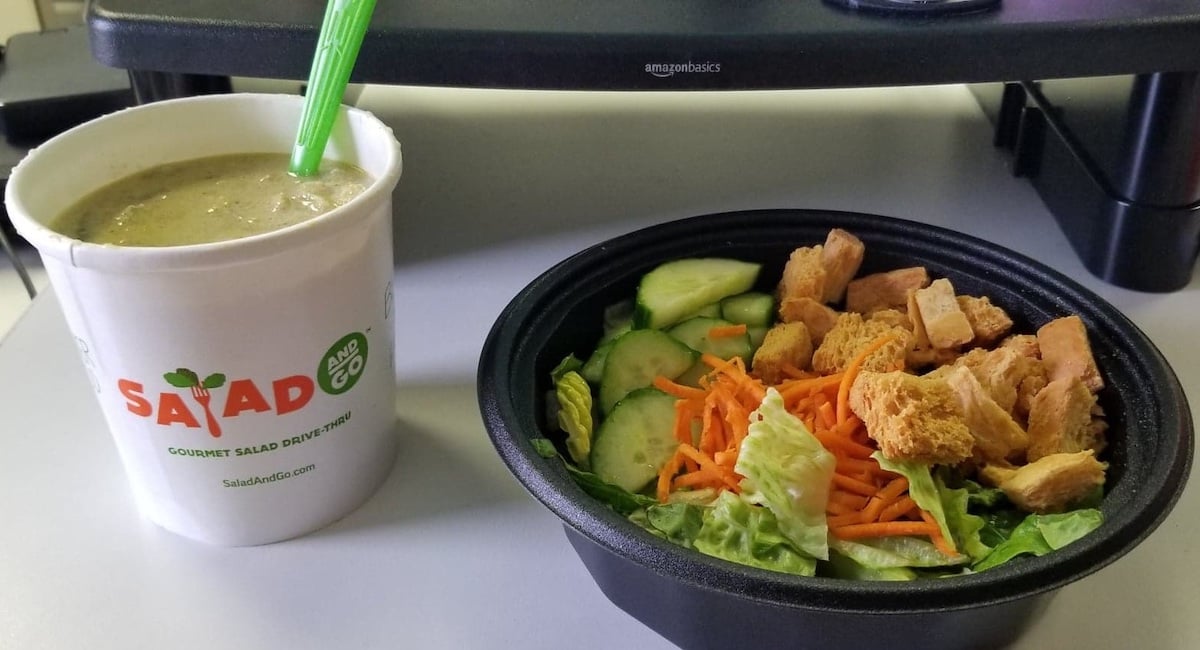A cup of green soup with a straw next to a black bowl containing a salad with lettuce, shredded carrots, cucumber slices, and croutons on a table.