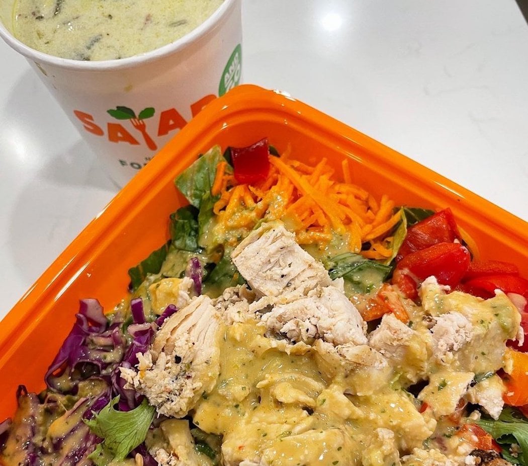 A colorful salad with grilled chicken, shredded carrots, red bell peppers, and purple cabbage in an orange container, topped with dressing. A cup of soup sits in the background on a white surface.