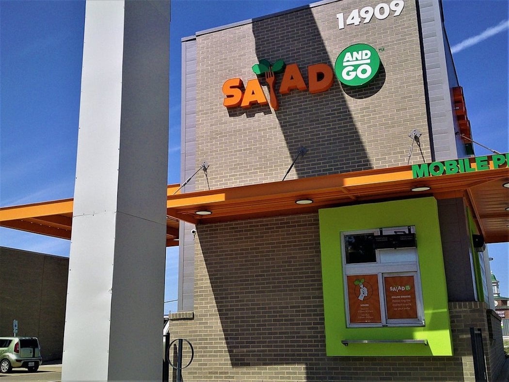 A Salad and Go restaurant with a drive-thru window is shown against a clear blue sky. The building is tan brick with orange and green signage and the address number 14909 displayed at the top.