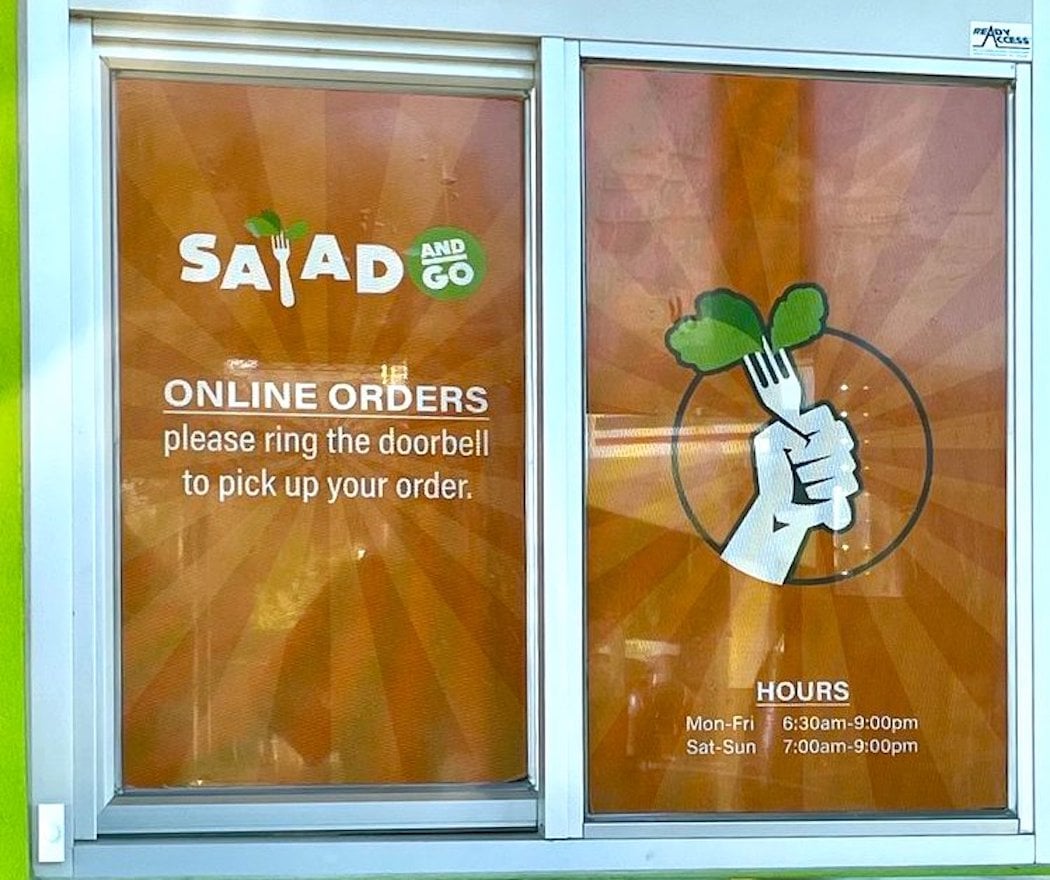 A Salad and Go window sign shows online order instructions to ring the doorbell for pickup, with business hours listed below and a graphic of a fist holding a fork with leafy greens.