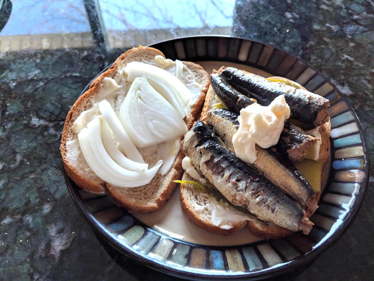 Two slices of bread on a plate, one topped with mayonnaise and raw onion slices, the other with sardines, pickles, and a dollop of mayonnaise. The plate rests on a dark countertop.