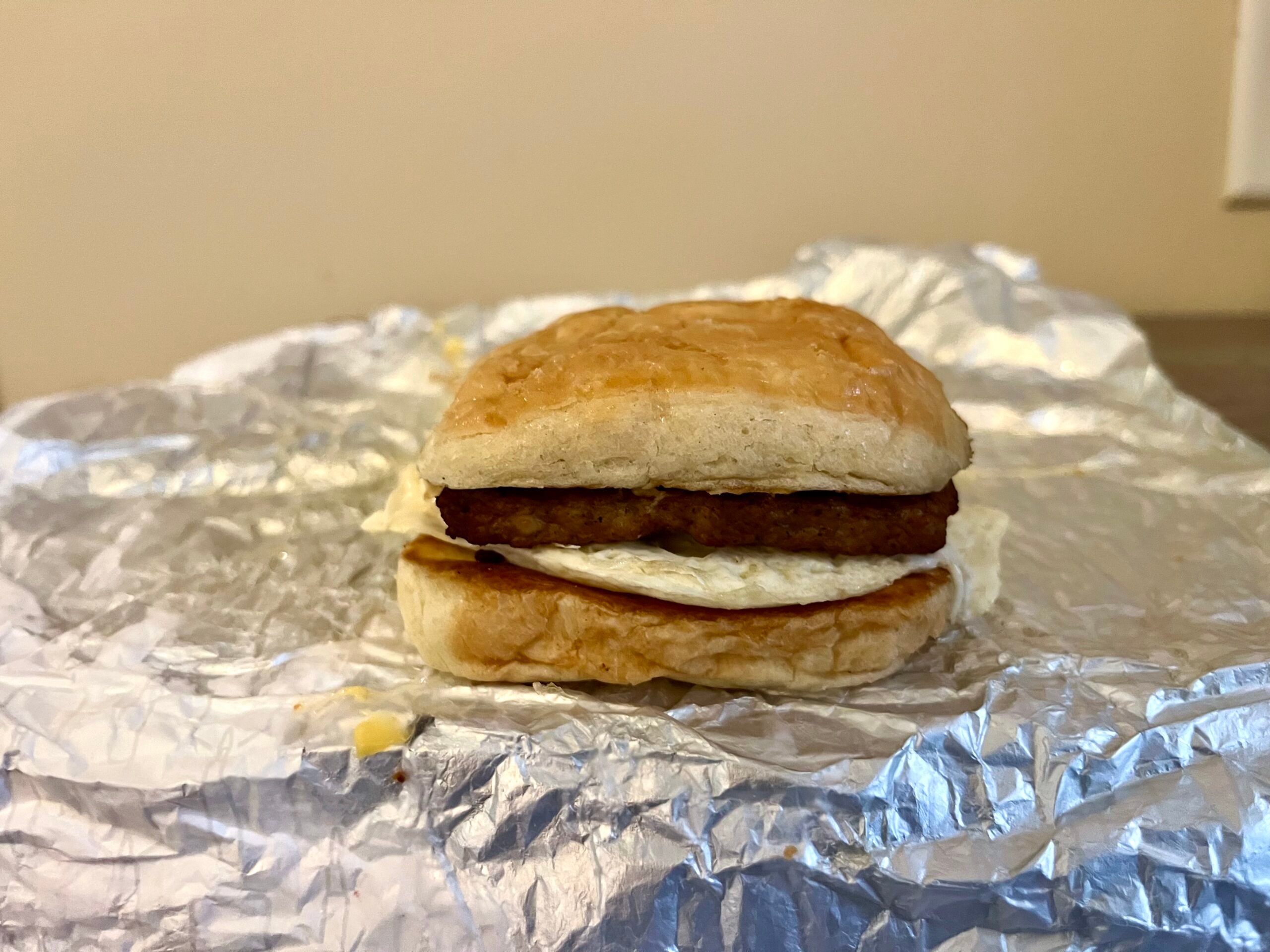 A breakfast sandwich with egg and sausage on a biscuit sits on crumpled aluminum foil with a plain beige background.