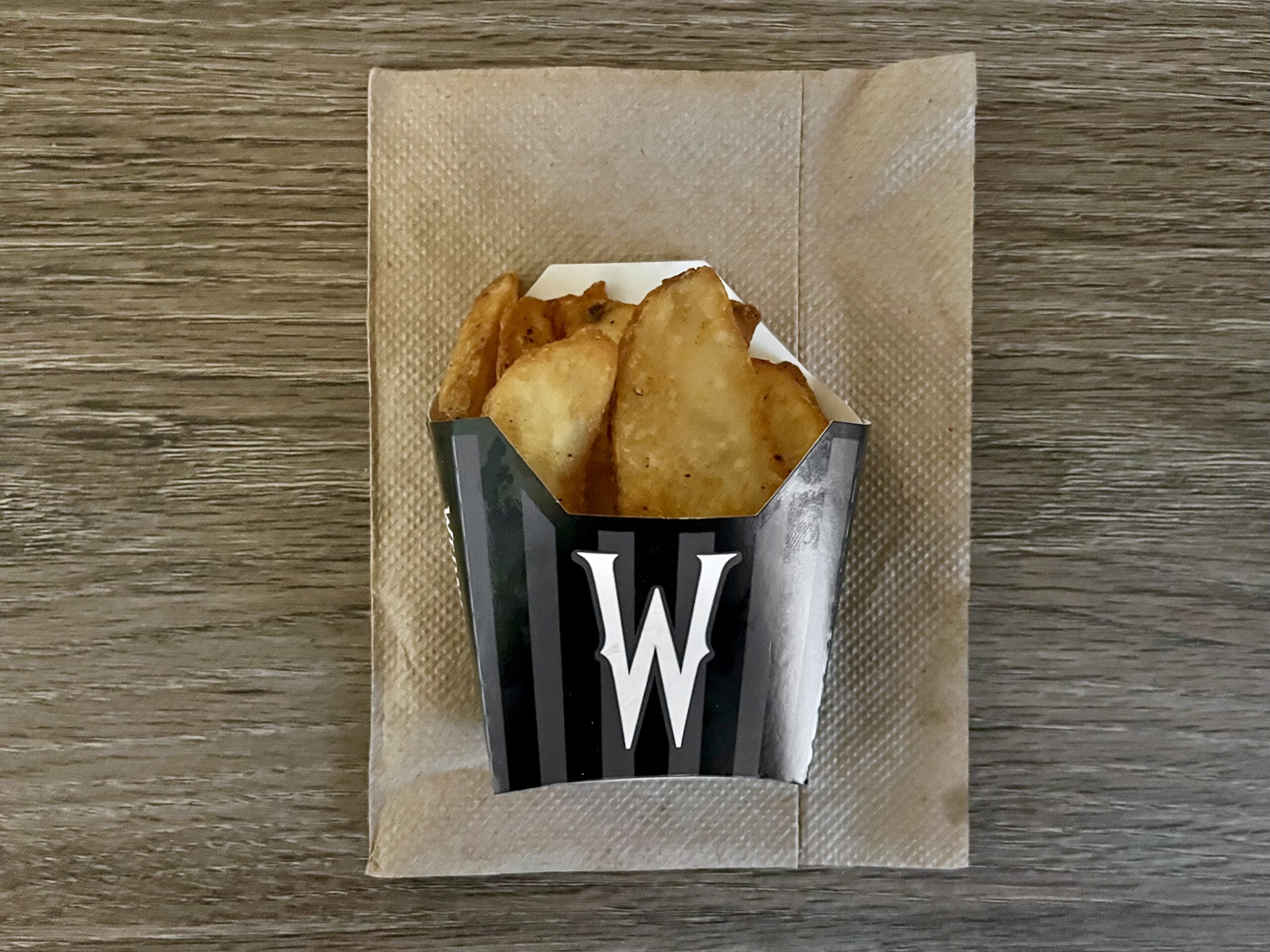 A black container with a white "W" logo holds seasoned potato wedges, placed on a brown napkin on a wooden surface.