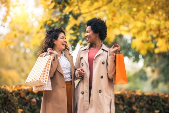 Two women wearing trench coats smile and talk while holding shopping bags outdoors. Autumn leaves and trees with yellow foliage create a warm, cheerful background.
