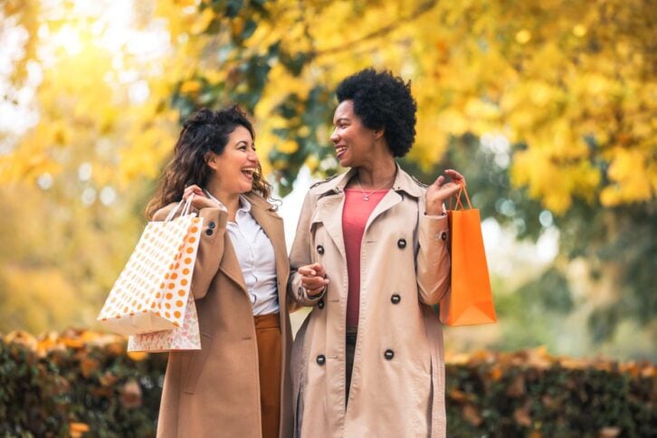 Two women wearing trench coats smile and talk while holding shopping bags outdoors. Autumn leaves and trees with yellow foliage create a warm, cheerful background.
