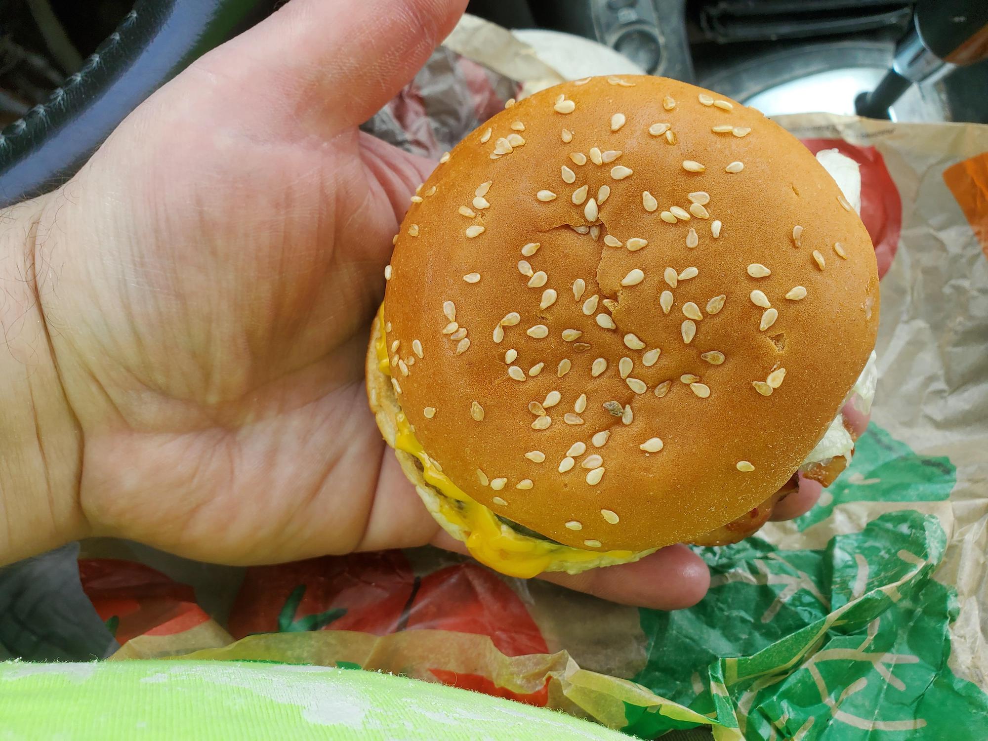 A hand holding a sesame seed hamburger with some mustard visible on the side, over a crumpled fast-food wrapper. Burger King Whopper