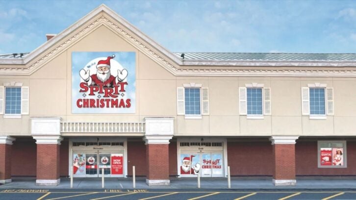 The front of a beige and red storefront with a large "Spirit Christmas" sign featuring Santa Claus. Festive windows hint at the magic found at Spirit Christmas locations. The clear blue sky shines above, welcoming you to Spirit Christmas 2025.