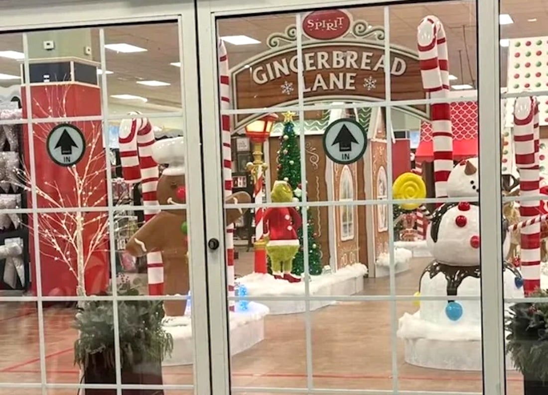 A store entrance decorated with giant candy canes, a gingerbread man, a snowman, and a festive “Gingerbread Lane” sign, viewed through glass doors with arrow “IN” signs. Holiday decorations fill the background.