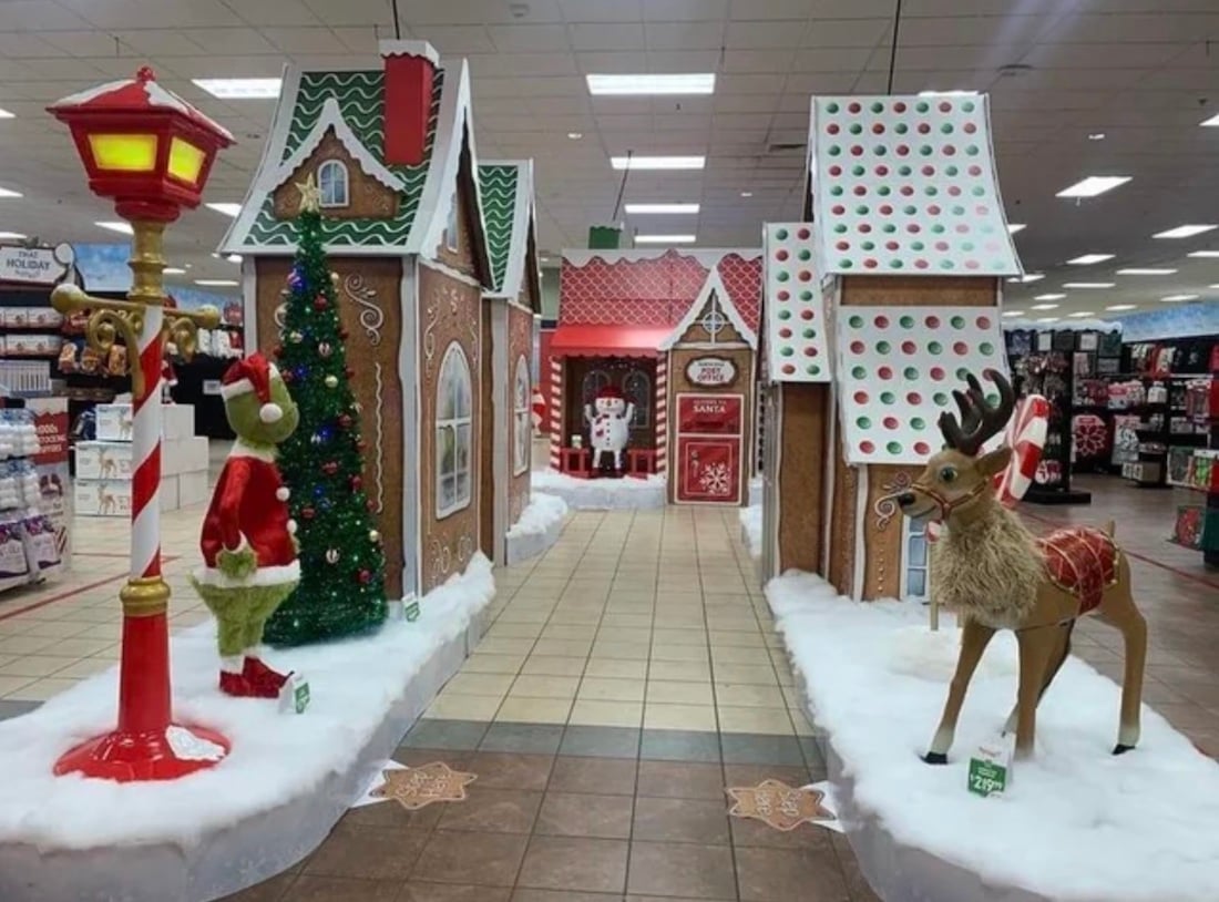 A festive store display captures the spirit of Christmas with gingerbread-style houses, a Christmas tree, the Grinch in a Santa suit, a large reindeer, fake snow, and Santa welcoming visitors at the end of a decorated pathway.