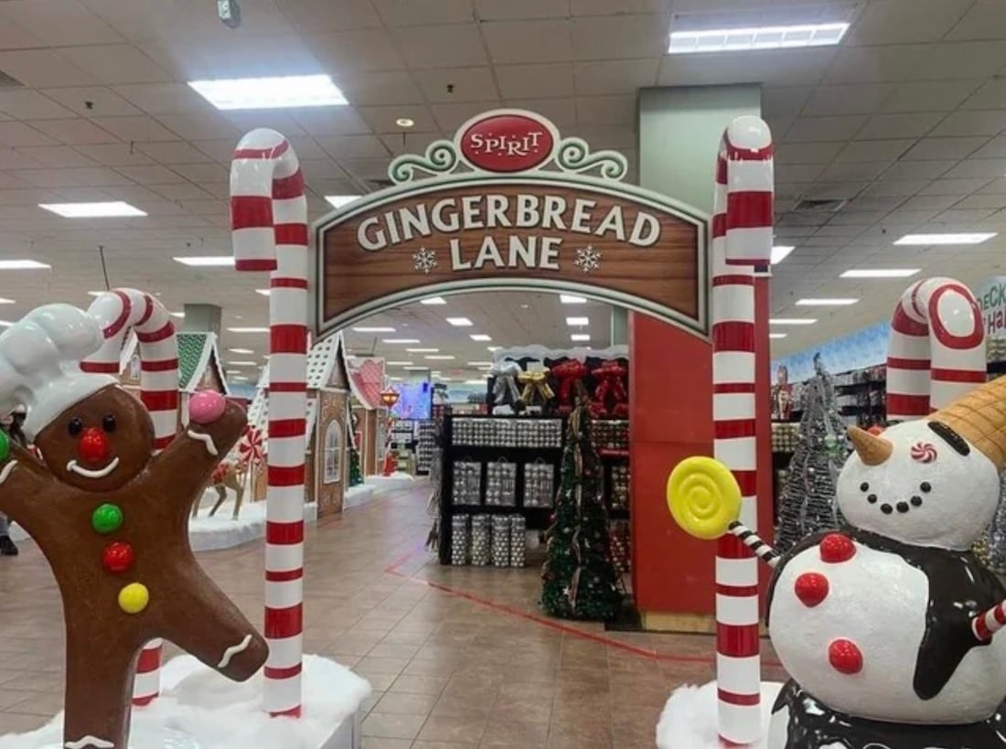A festive store display with a "Gingerbread Lane" sign, candy cane poles, a smiling gingerbread figure, and a cheerful snowman holding a yellow lollipop captures the spirit of Christmas. Holiday decorations fill the background.