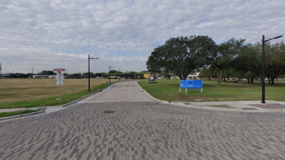 A paved road lined with streetlights runs through an open grassy area with trees, a blue sign on the right, and a cloudy sky overhead. There are few cars and buildings in the distant background.