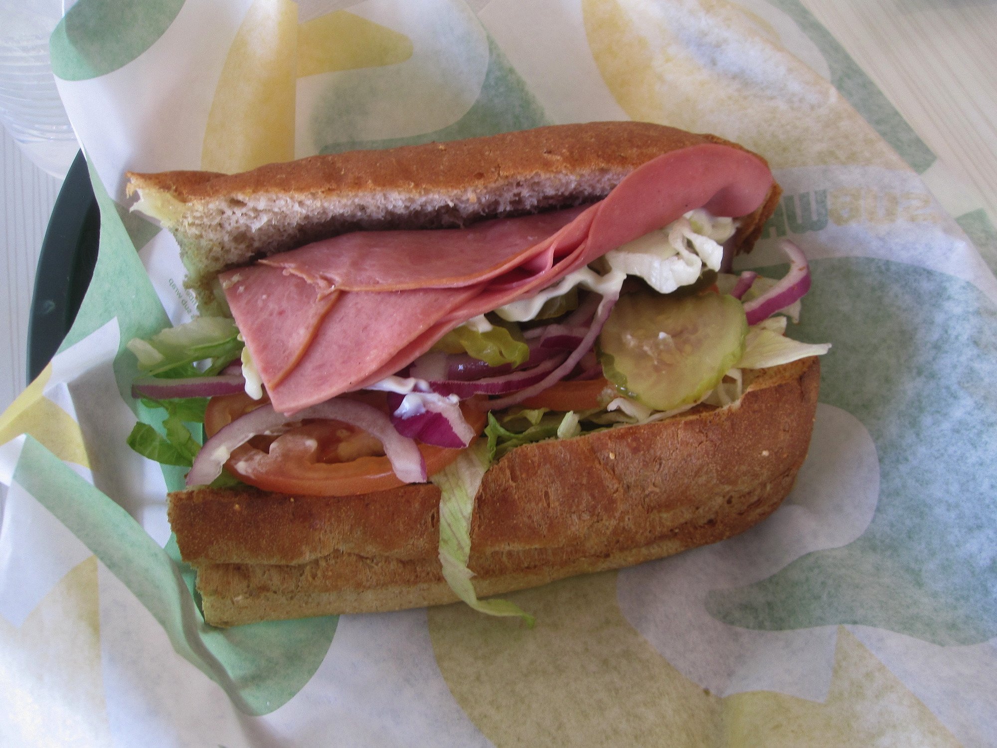 A sub sandwich on whole wheat bread with layers of ham, lettuce, tomato, red onion, and pickles, served on Subway branded sandwich paper.