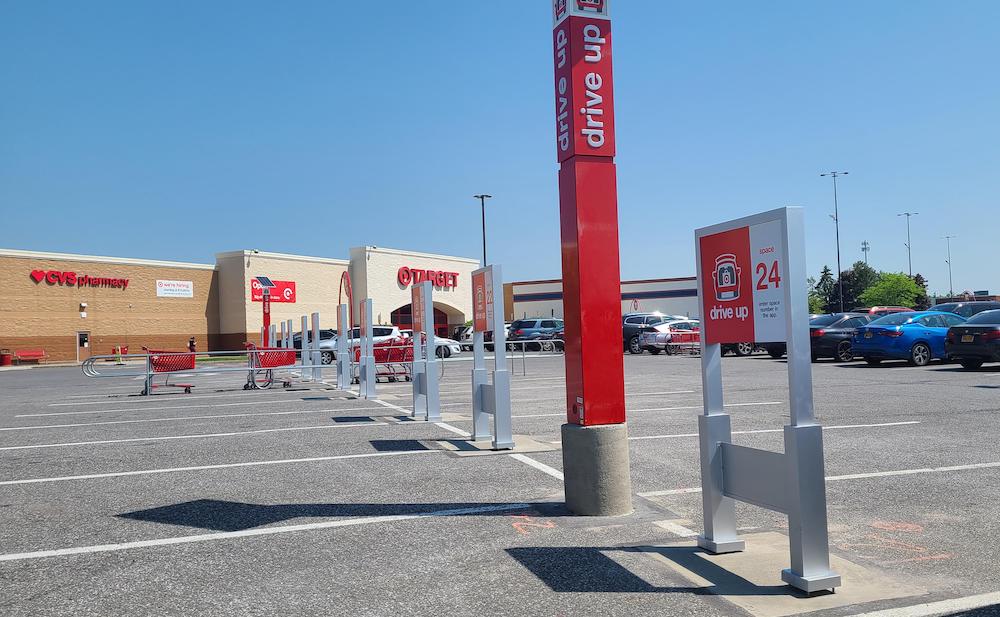 A Target parking lot with designated "Drive Up" pickup spots, a tall red sign, parked cars, and shopping carts near the store entrance on a sunny day. Cover image for new Target stores article.