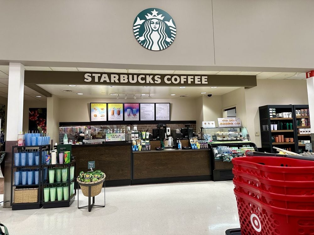 A Starbucks Coffee counter inside a Target store, with a display of drinks and merchandise in front and red Target shopping carts to the right. The Starbucks logo is visible above the counter.