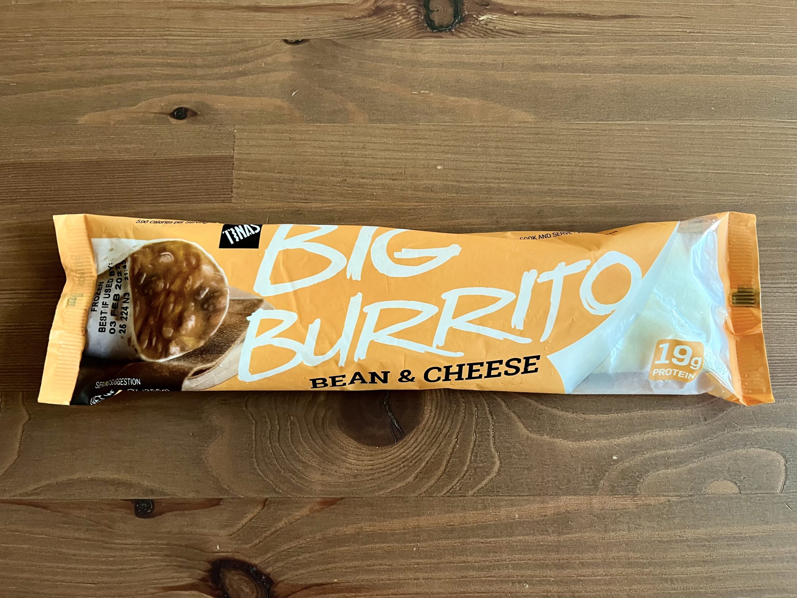 A packaged bean and cheese Big Burrito on a wooden surface. The orange and white wrapper highlights "Big Burrito" in large text and "19g protein" in a yellow circle.