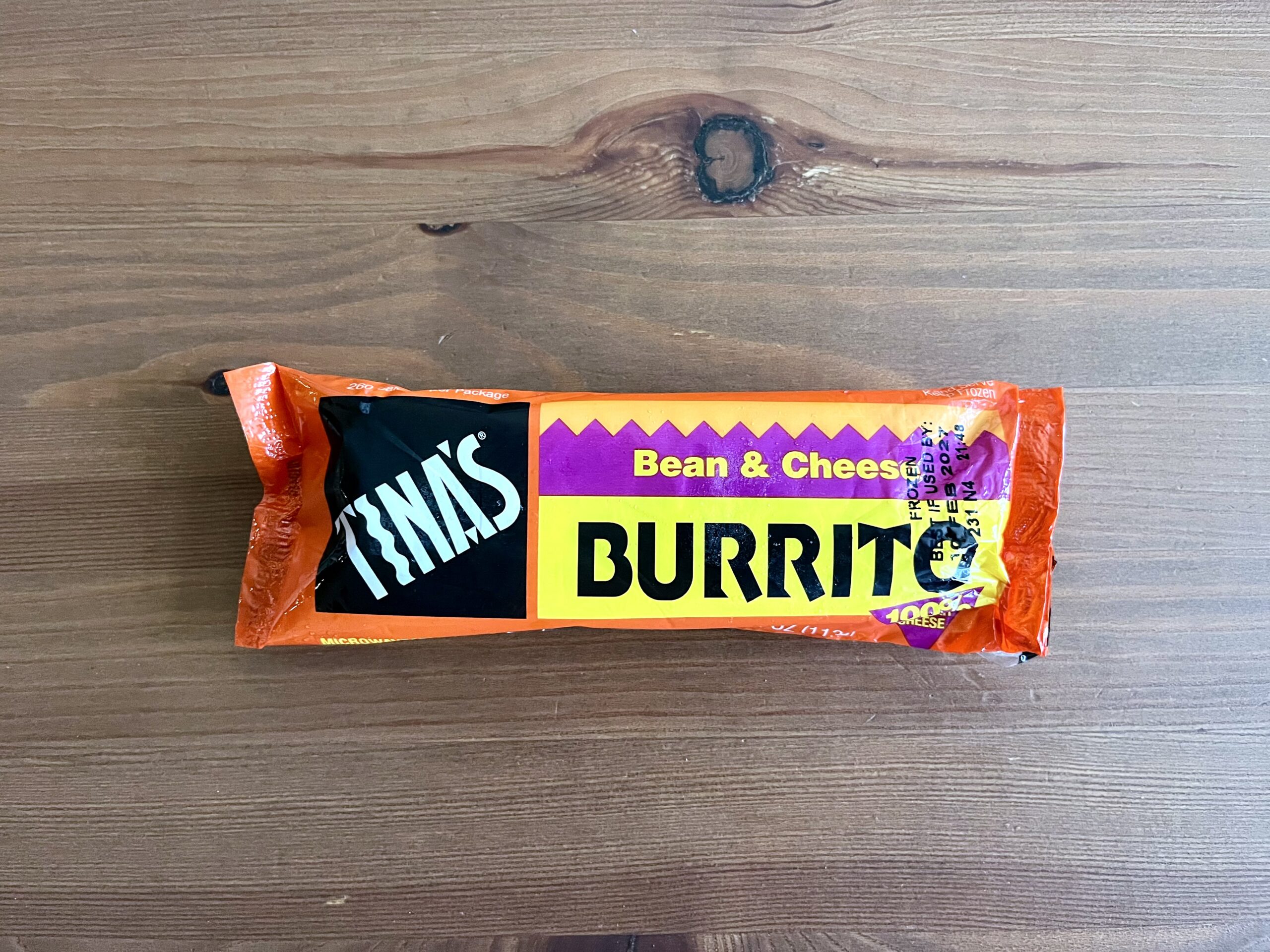 A packaged Tina’s Bean & Cheese Burrito sits on a wooden surface. The wrapper is mostly orange with yellow and purple accents and bold black and white lettering.