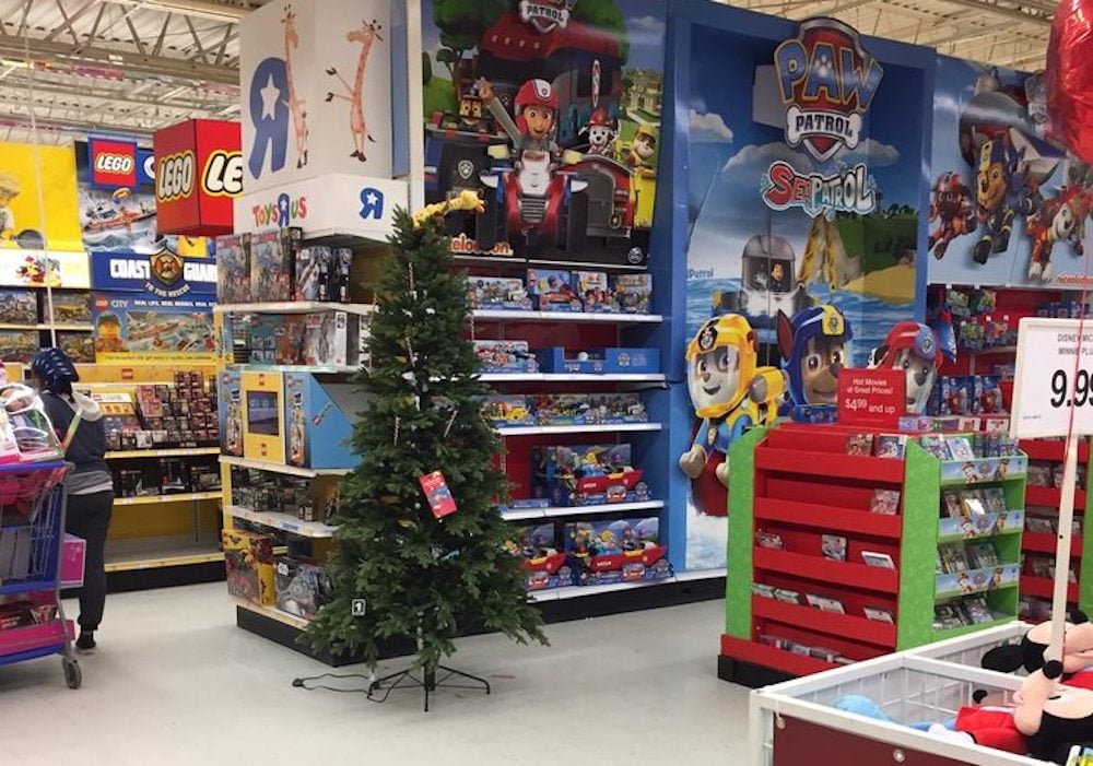 A partially decorated Christmas tree stands in the aisle of a toy store filled with LEGO, Paw Patrol, and various toys. Colorful shelves and large character posters cover the walls. Shoppers browse nearby.