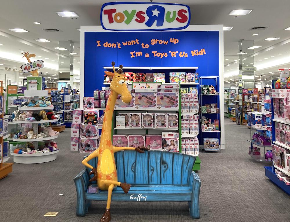 A Geoffrey the Giraffe statue sits on a blue bench in front of a Toys "R" Us display, surrounded by shelves of toys. A sign above reads, "I don’t want to grow up, I’m a Toys ‘R’ Us kid!