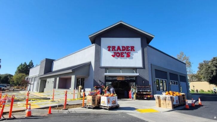 A Trader Joe's store exterior on a sunny day, with the logo above the entrance. People are entering and exiting, and pumpkins and other items are displayed outside. Orange cones and caution tape line the sidewalk.