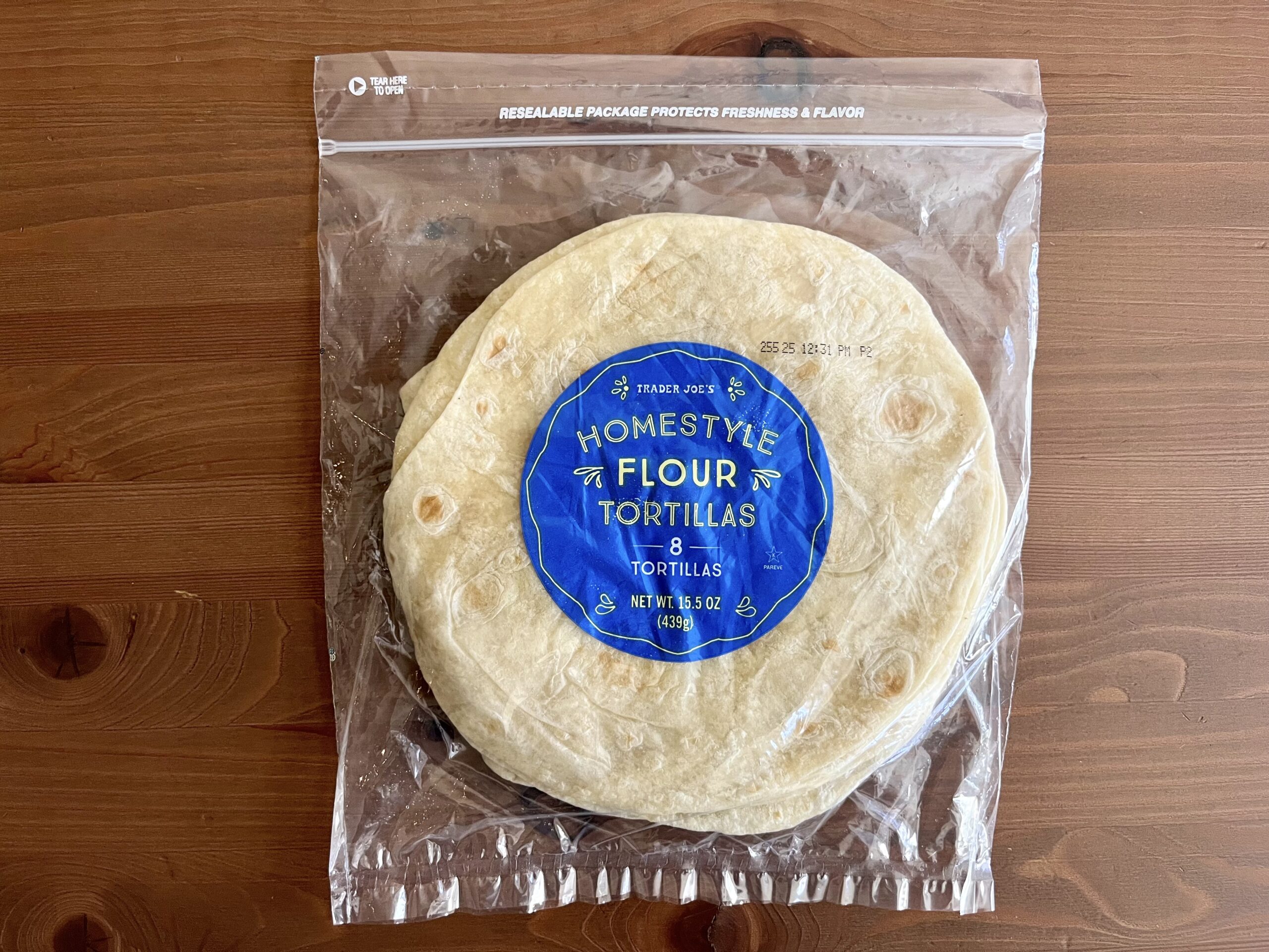 A package of Trader Joe’s Homestyle Flour Tortillas sits on a wooden surface. The clear resealable bag contains eight tortillas with a blue label listing the product name and weight.