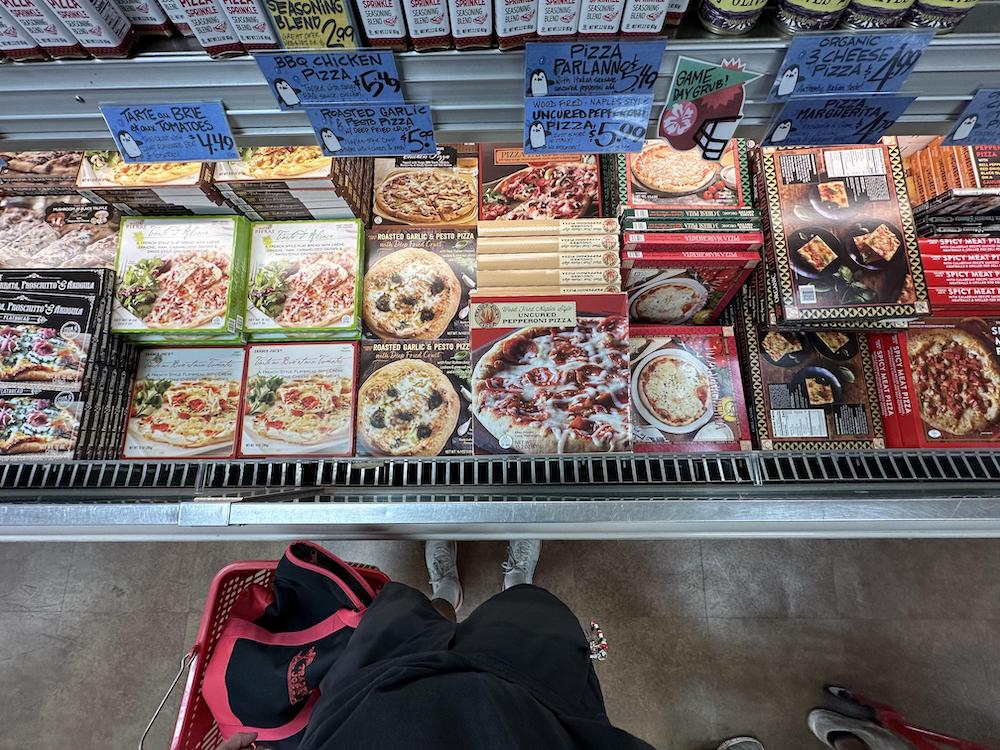 A person stands in front of a freezer stocked with various frozen pizzas and Italian meals at a grocery store. Shopping basket and handwritten price tags are visible. View is from the shopper's perspective, looking down.