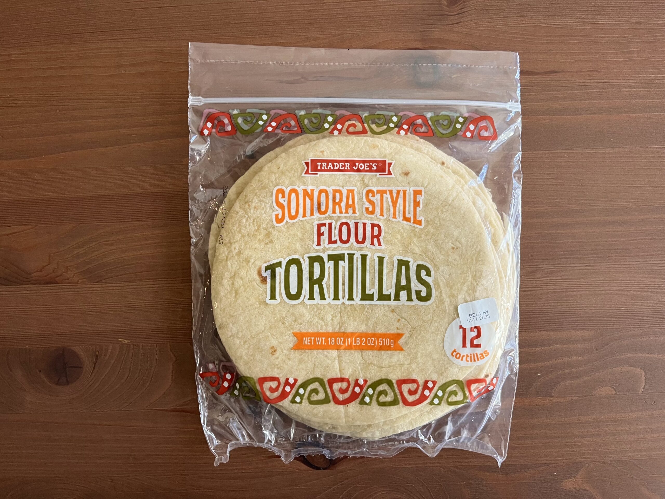 A package of Trader Joe’s Sonora Style Flour Tortillas sits on a wooden surface. The clear plastic bag displays 12 tortillas and colorful decorative patterns along the top and bottom edges.