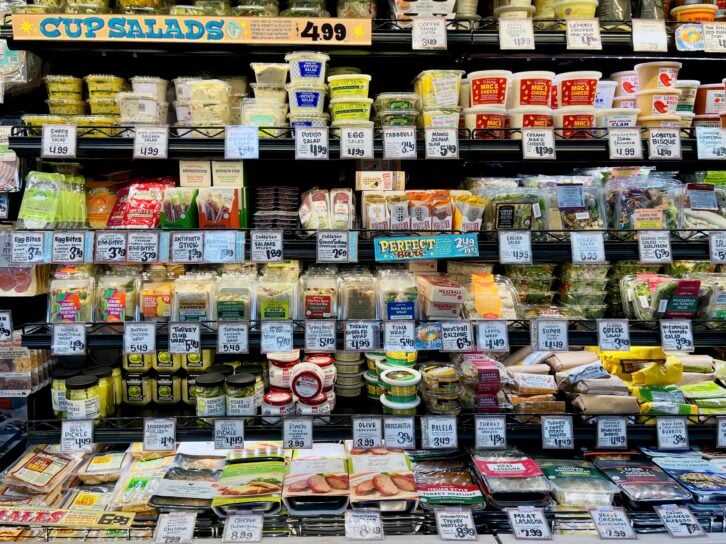 A well-stocked grocery store shelf displays various packaged salads, dips, cheeses, and deli items, with labeled price tags and a sign reading “CUP SALADS $4.99” at the top.