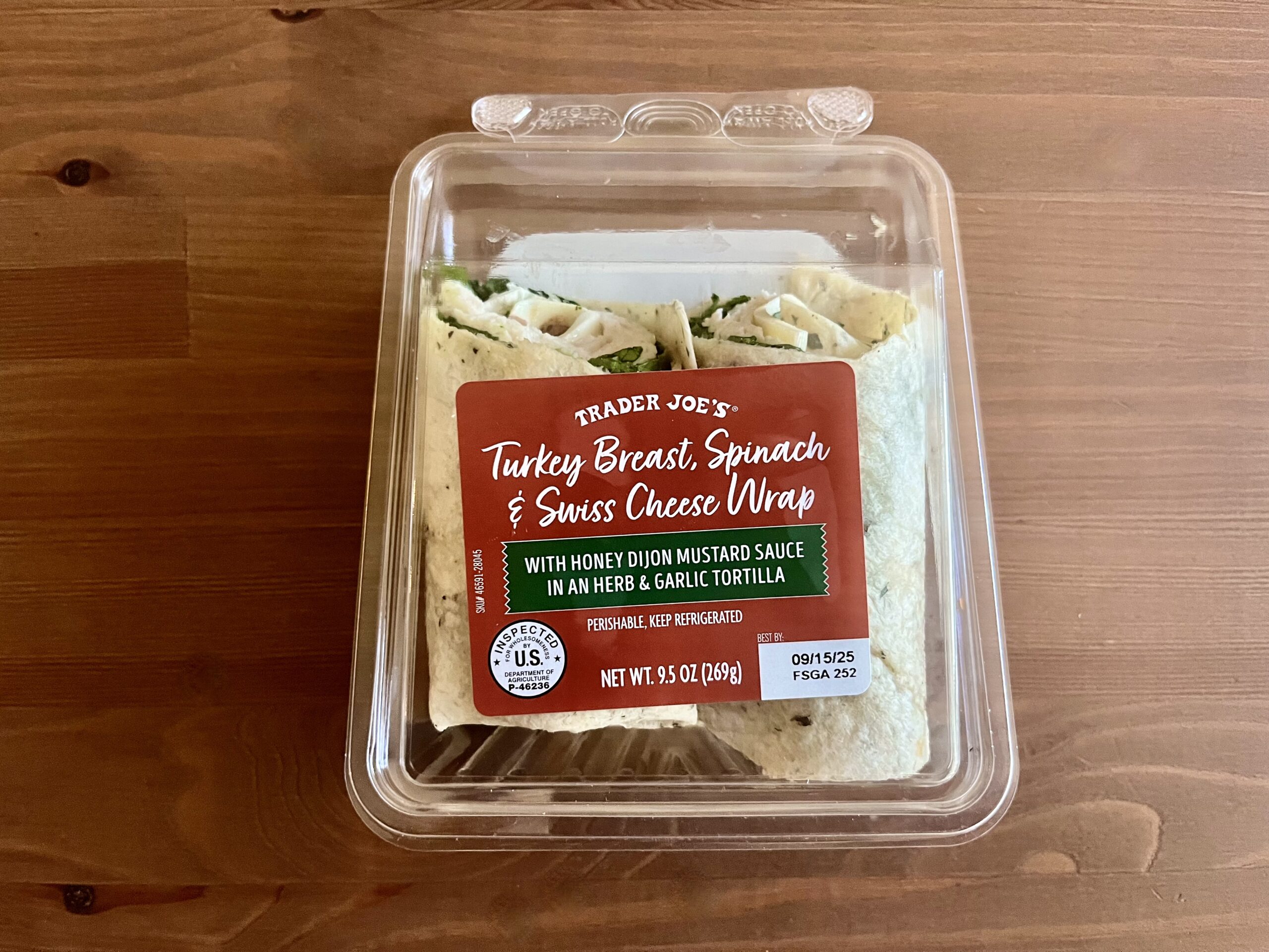 A clear plastic container holds Trader Joe’s Turkey Breast, Spinach & Swiss Cheese Wrap with honey dijon mustard sauce. The label is red and green, and the wraps are visible inside, resting on a wooden surface.