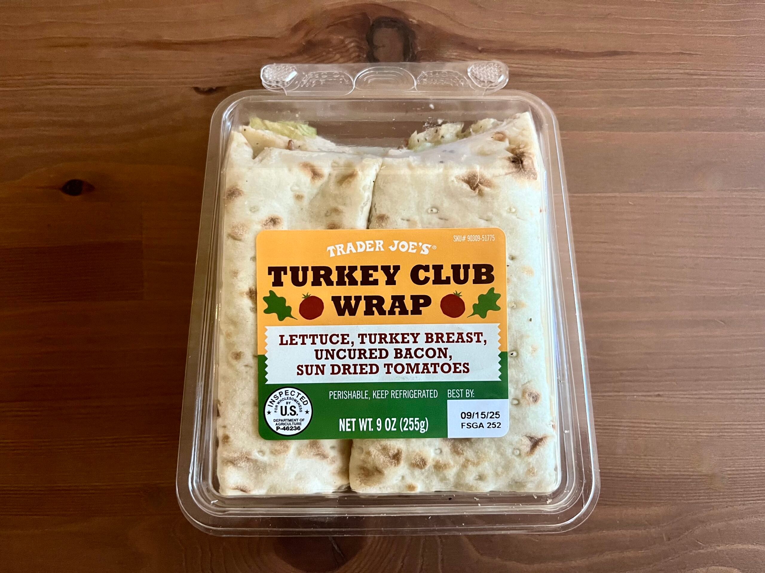 A plastic container of Trader Joe’s Turkey Club Wrap sits on a wooden table. The label lists ingredients: lettuce, turkey breast, uncured bacon, and sun-dried tomatoes. The wrap is partially visible through the clear lid.