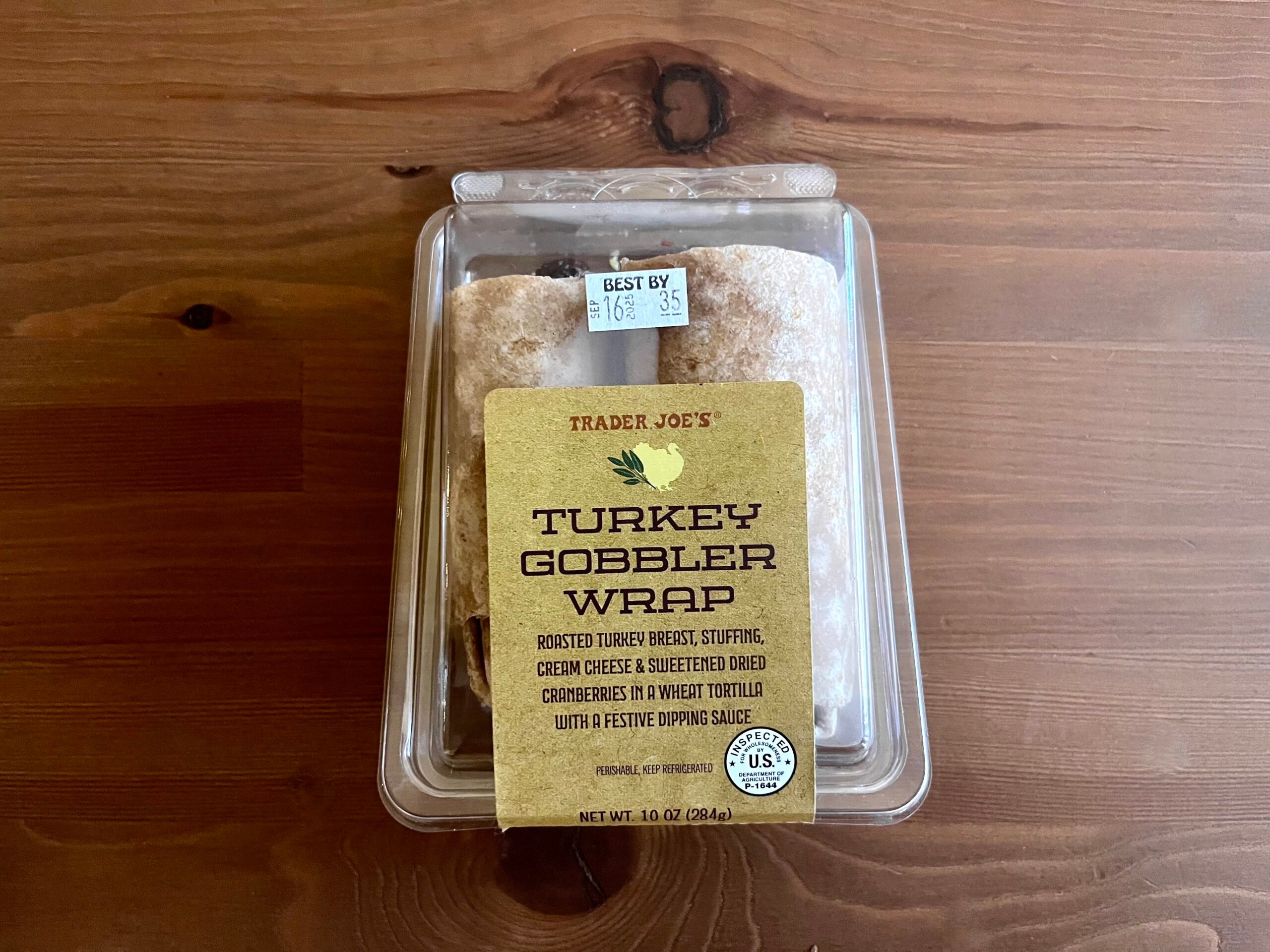 A plastic container of Trader Joe's Turkey Gobbler Wrap sits on a wooden surface. The label lists ingredients: turkey breast, stuffing, cream cheese, sweetened dried cranberries, and a dipping sauce.
