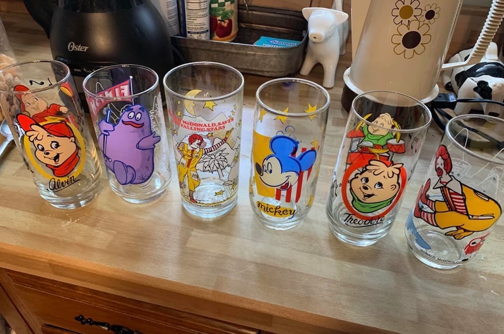 Six vintage collectible drinking glasses featuring colorful cartoon characters, including Alvin, Grimace, Ronald McDonald, Mickey Mouse, Theodore, and Ronald McDonald riding a hamburger, lined up on a wooden kitchen counter.