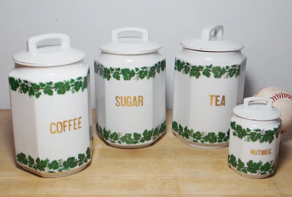 Four white ceramic canisters labeled coffee, sugar, tea, and nutmeg with green ivy patterns sit on a wooden surface; a baseball is in the background.