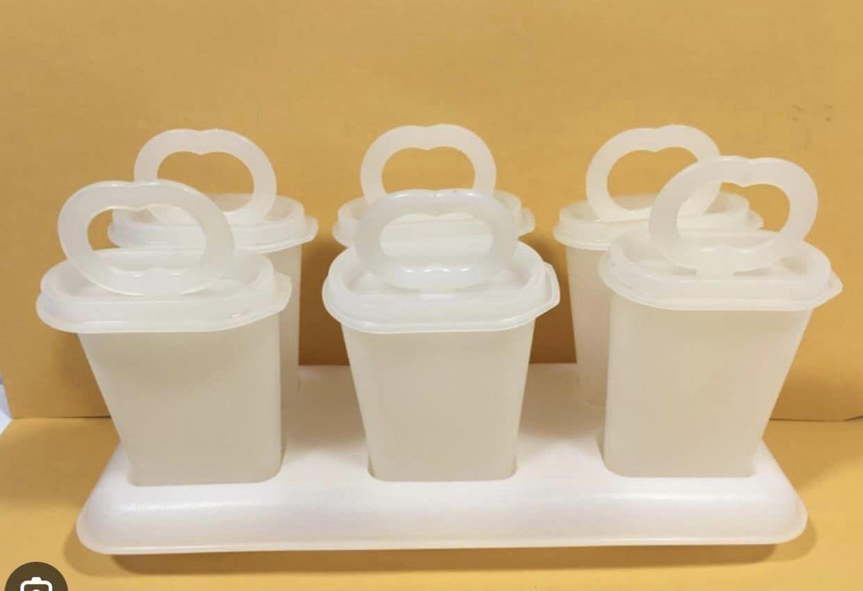 Five white vintage Tupperware plastic popsicle molds with oval handles are arranged on a rectangular white tray, set against a plain yellow background.